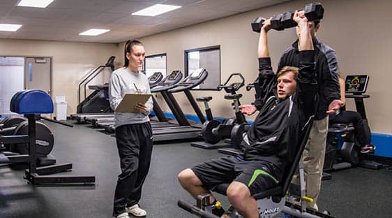 Three students in the FLCC Fitness Center. One student is documenting a student participant as he lifts free weights