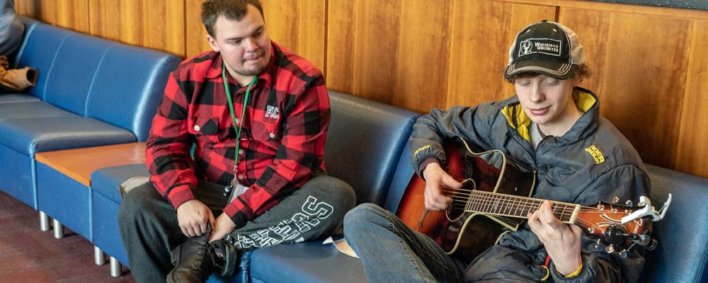 Student playing guitar in the lounge area as a student listens.