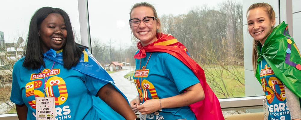 Three smiling college students. Each wears a shirt celebrating the 50th anniversary of Sesame Street and a colorful, homemade cape. 