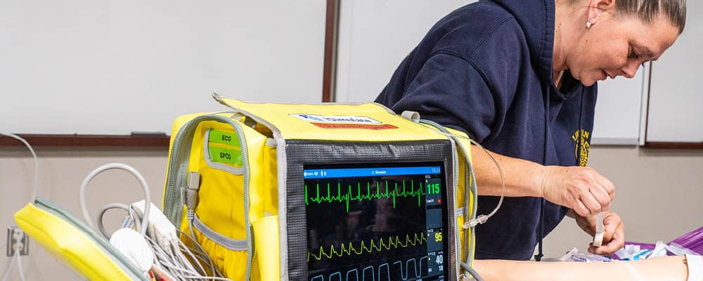 An EMT - Paramedic student preparing emergency medical supplies. A screen in the foreground displays vitals.