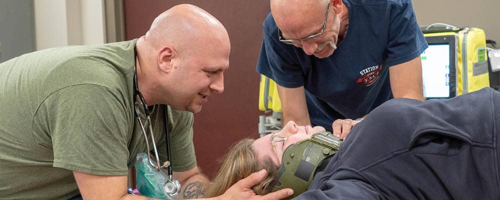 Two Emergency Medical Technician instructors demonstrate how to secure a brace on an injured woman's neck.