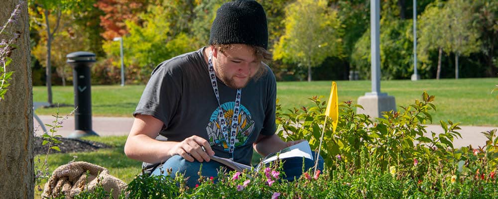 A college student sits cross-legged in the grass studying nearby plants and flowers. In the distance, trees are beginning to take on their autumn hues.